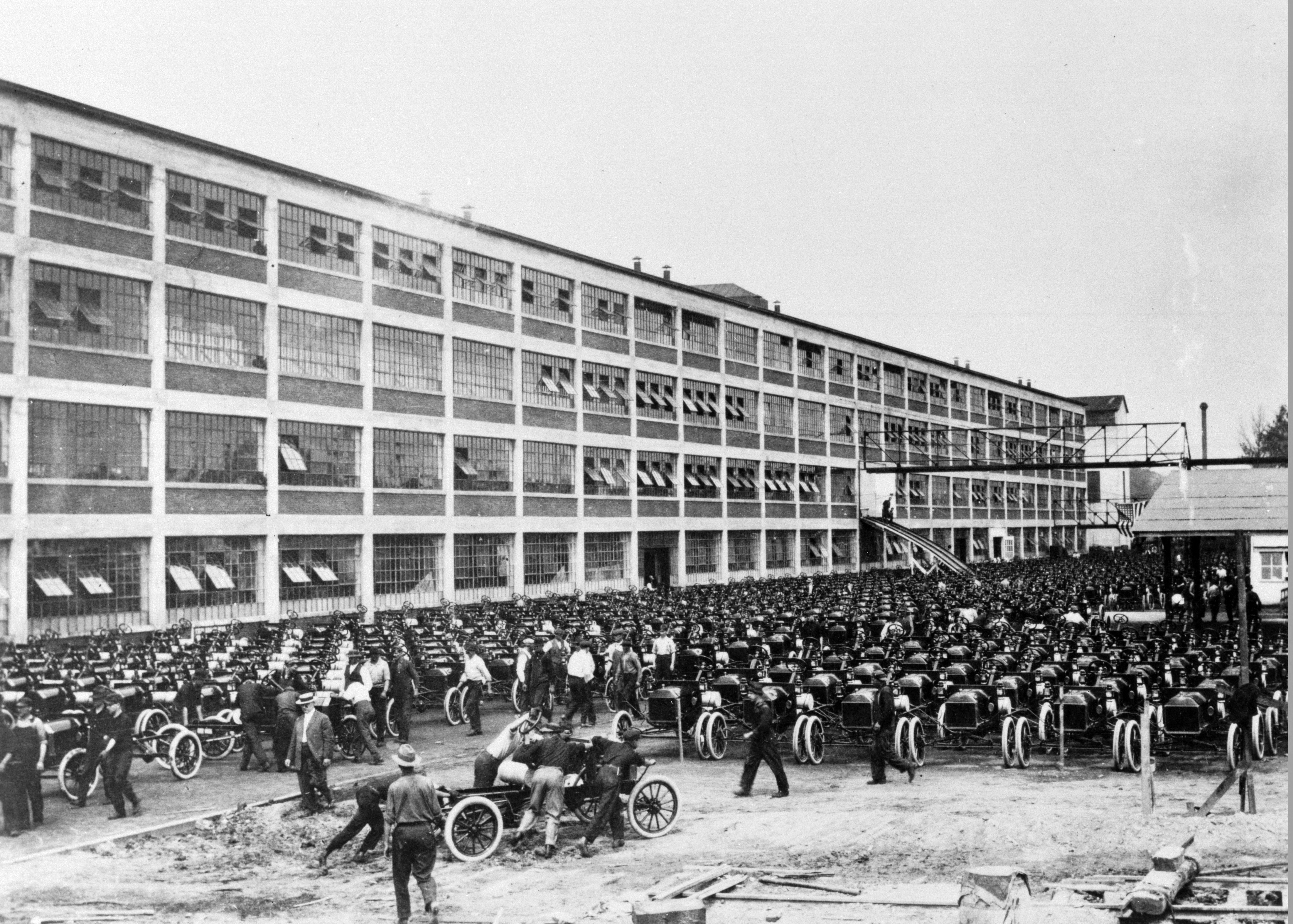 A black and white photo shows a lot full of old-school vehicles next to a tall building with many windows.